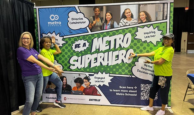 alt= metro teachers and battelle employees posing in front of our be a metro superhero banner